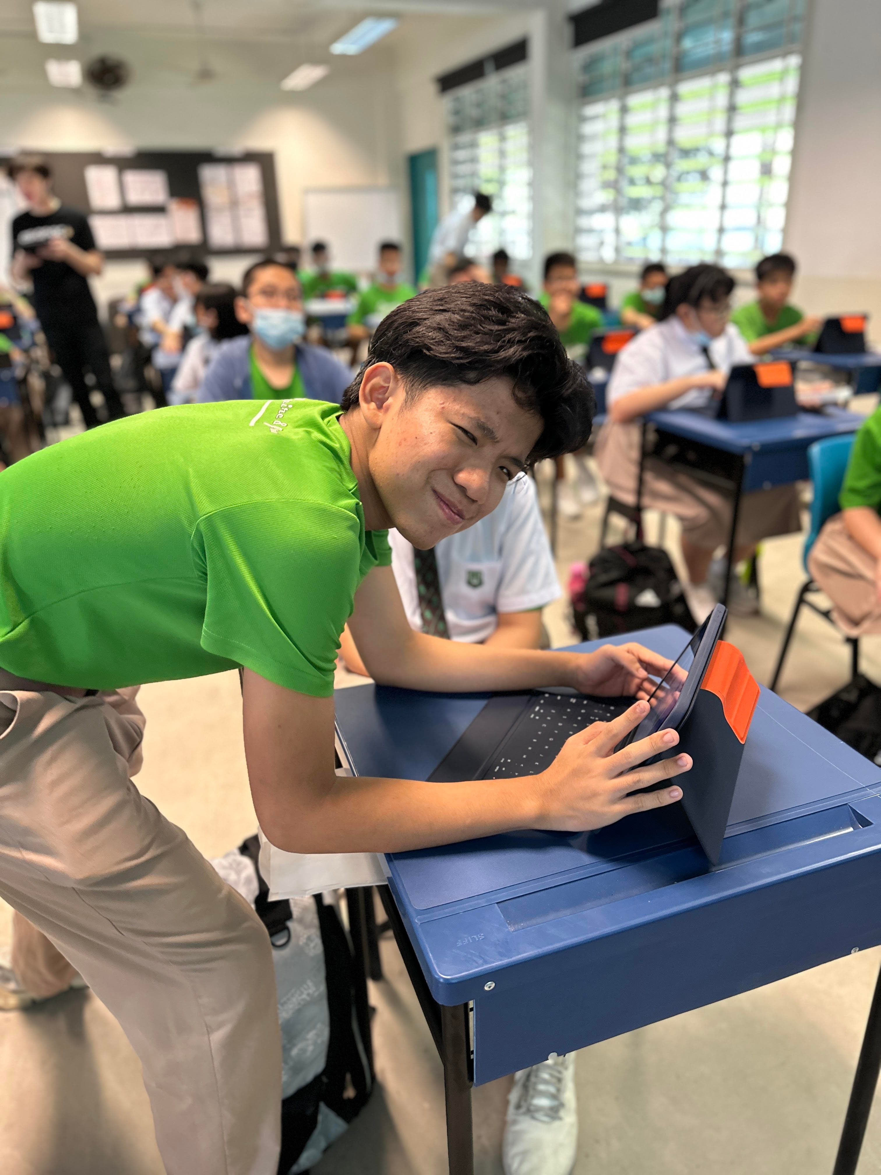 A smiling teen in a green shirt leans over a blue desk using a tablet in a busy classroom.
